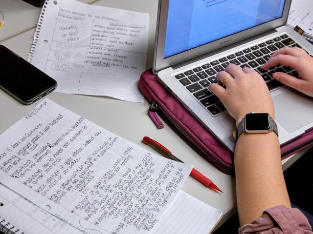 hands typing on a laptop with a handwritten paper on the table.